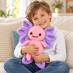 Puede incluir: Un peluche de ajolote hecho de hilo suave y texturizado. La criatura presenta un cuerpo redondo de color rosa claro con brazos y piernas pequeños, y grandes branquias plumosas de color morado vibrante a los lados de la cabeza.