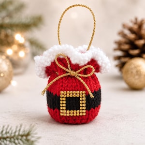 Op de afbeelding: Een klein rood en zwart gebreid trekkoordzakje met een witte pluizige rand aan de bovenkant, een gouden koordhandvat en een gouden strik. Een vierkante gesp versierd met kleine gouden strass-steentjes zit op de zwarte band.