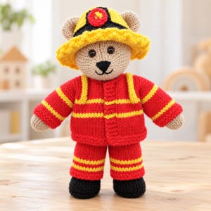 May include: A hand-knitted teddy bear dressed in a red firefighter uniform with yellow stripes. The bear wears a matching yellow and black firefighter helmet, has a beige body, black stitched nose and mouth, and black knitted boots on its feet.