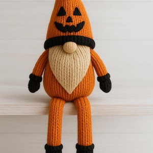 May include: A knitted Halloween gnome doll in orange, black, and beige. The gnome has a pumpkin-shaped head with a carved face, a black brim, and a beige beard. It is wearing black gloves and boots, and is sitting on a white shelf.