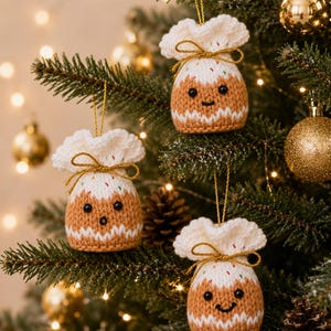 Op de afbeelding: Drie gebreide kerstpudding-ornamenten hangen aan dennentakken. Elk ornament heeft een bruine basis, een witte bovenkant met kleurrijke spikkels, een witte gegolfde rand, een gouden ophanglus en een gouden strik met geborduurde gezichtjes.