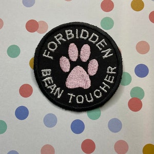 Könnte beinhalten: Ein gestickter schwarzer und weißer Aufnäher mit einem rosa Pfotenabdruck in der Mitte. Der Text um den Pfotenabdruck lautet "Forbidden Bean Toucher".