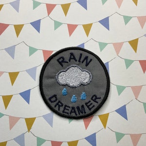 May include: A grey round iron-on patch with the text "RAIN DREAMER" in navy blue embroidery. A white embroidered cloud with three blue raindrops is centred on the patch.