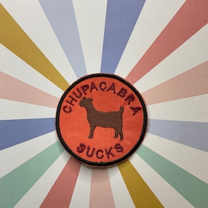 May include: A round, orange embroidered patch with a black outline. The patch features a brown silhouette of a goat and the text "Chupacabra Sucks" in maroon.
