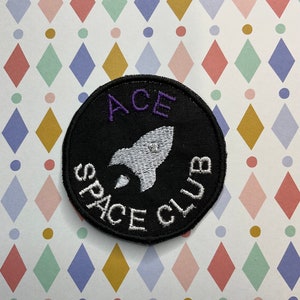 Könnte beinhalten: Ein schwarzer gestickter Aufnäher mit einer weißen Rakete und dem Text "ACE SPACE CLUB" in Weiß mit "ACE" in Lila.