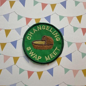 May include: A green embroidered patch with the text "CHANGELING SWAP MEET" in a circle. The patch features a brown baby crib with a yellow blanket inside.