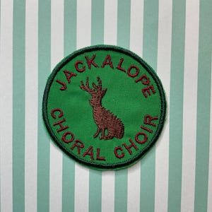 May include: A green circular patch with red embroidered text that reads "Jackalope Choral Choir". A brown embroidered jackalope with antlers is in the centre of the patch.