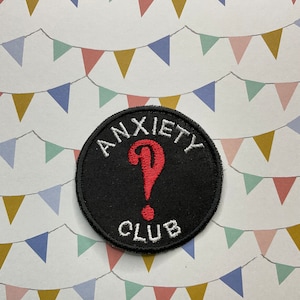May include: Black embroidered patch with white text that reads "Anxiety Club" and a red question mark. The patch is on a white background with a pattern of colourful bunting.