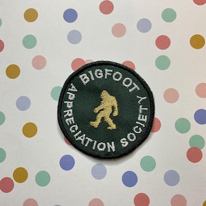 May include: An embroidered patch in green with white text that reads "Bigfoot Appreciation Society". A gold silhouette of Bigfoot walking is in the centre of the patch.