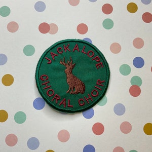 May include: A green circular patch with red embroidered text that reads "Jackalope Choral Choir". A brown embroidered jackalope is in the centre of the patch.