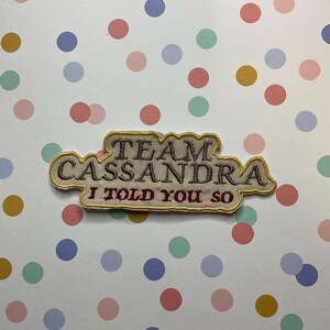 Könnte beinhalten: Ein beigefarbener gestickter Aufnäher mit dem Text "Team Cassandra I told you so" in braunem und goldenem Garn.