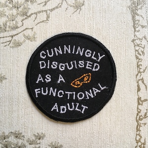 May include: Black round patch with white embroidery that reads "Cunningly Disguised as a Functional Adult". A small orange graphic of a pair of glasses is between the words "as a" and "functional".
