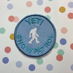 May include: A blue and white embroidered patch with the text "YETI SNOW PATROL" in turquoise thread. A white silhouette of a Yeti is in the centre of the patch.