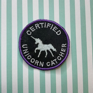 May include: A round, black patch with a purple border. The patch has white embroidery that reads "CERTIFIED UNICORN CATCHER" and features a white unicorn silhouette.