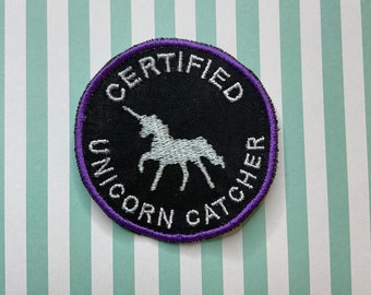 May include: A round, black patch with a purple border. The patch has white embroidery that reads "CERTIFIED UNICORN CATCHER" and features a white unicorn silhouette.