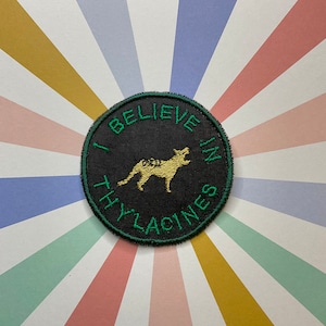 May include: A circular black patch with green embroidered text: "I BELIEVE IN THYLACINES." The patch features a yellow embroidered thylacine. The background is a pattern of radiating stripes in various pastel colours.
