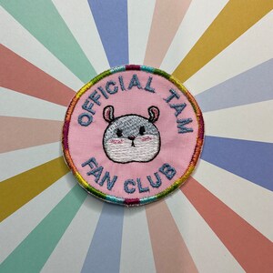 May include: A pink embroidered patch with a blue and grey hamster in the centre. The text around the hamster reads "Official Fan Club".