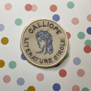 May include: A beige embroidered patch with the text "Calliope Literature Circle" in brown thread. A blue embroidered image of a woman's head is in the centre of the patch.