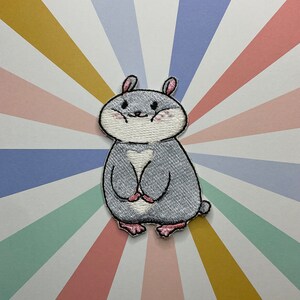 以下が含まれることがあります： 灰色の毛並み、ピンクの耳、バラ色の頬をした漫画のハムスターの刺繍パッチ。 ハムスターは白いハート型の胸をしており、前足を合わせています。 パッチは、カラフルな放射状のストライプの背景に設定されています。