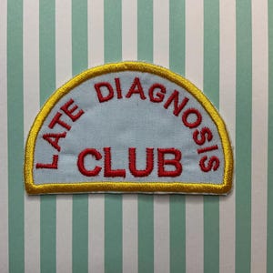 May include: A light blue patch with the words "LATE DIAGNOSIS CLUB" embroidered in red. The patch has a yellow border and is set against a striped background.