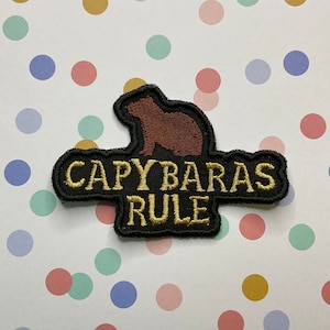 Peut inclure: Un patch brodé noir et or avec une silhouette de capybara marron et le texte "Capybaras Rule".