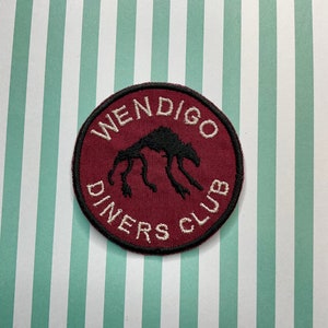 May include: A maroon circular patch with white embroidered text that reads "Wendigo Diners Club". A black silhouette of a creature with multiple legs is in the centre of the patch.