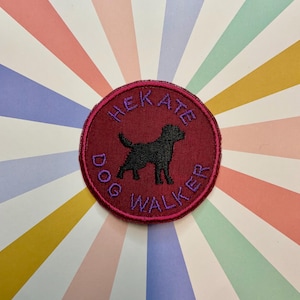 May include: A circular embroidered patch with a deep red background and a dark purple stitched border. The patch features a black silhouette of a dog in the center, surrounded by the purple embroidered text "HEKATE DOG WALKER" in a circular arc.