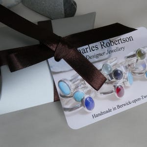 K&ouml;nnte beinhalten: Hellblaue Geschenkbox mit braunem Satinband. Ein Etikett zeigt Ringe mit farbigen Edelsteinen. Das Etikett tr&auml;gt die Aufschrift "Charles Robertson Designer Jewellery" und "Handmade in Berwick-upon-Tweed".