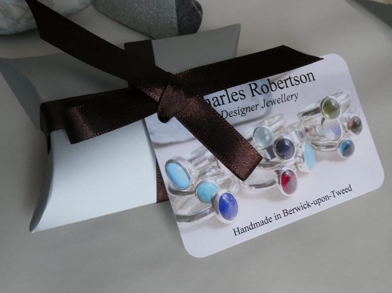 K&ouml;nnte beinhalten: Hellblaue Geschenkbox mit braunem Satinband. Ein Etikett zeigt Ringe mit farbigen Edelsteinen. Das Etikett tr&auml;gt die Aufschrift "Charles Robertson Designer Jewellery" und "Handmade in Berwick-upon-Tweed".