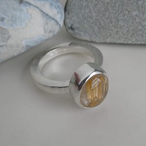 May include: A silver ring with a round, clear gemstone with gold inclusions. The gemstone is set in a silver bezel.