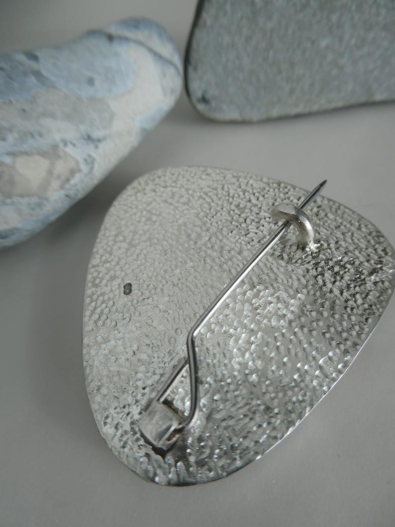 K&ouml;nnte beinhalten: Silberfarbene Brosche mit strukturierter, gehammerter Oberfl&auml;che. Die Brosche hat eine dreieckige Form mit abgerundeten Kanten und einem Nadelverschluss auf der R&uuml;ckseite. Zwei weitere graue, strukturierte Objekte sind zu sehen.