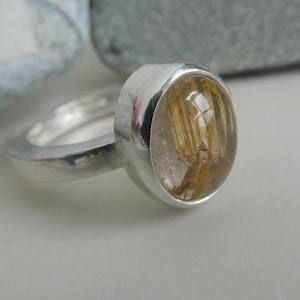 May include: A silver ring with a clear oval-shaped stone containing gold flecks. The stone is set in a bezel setting.