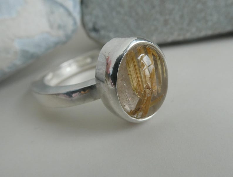 May include: A silver ring with a clear oval-shaped stone containing gold flecks. The stone is set in a bezel setting.