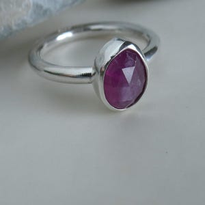 Peut inclure: Une bague en argent avec une pierre précieuse rose foncé, taillée en forme ovale et à facettes. La bague a un anneau lisse et poli, et la pierre est sertie dans un sertissage à lunette. La bague est présentée sur une surface claire.