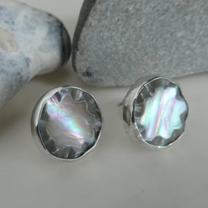 Op de afbeelding: Een paar zilveren oorbellen met iriserende abalone-schelpincrustaties. De oorbellen zijn rond en hebben een getextureerde rand.