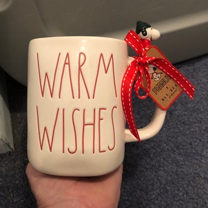 May include: White ceramic mug with red lettering that reads "warm wishes". The mug has a red ribbon and a small ceramic Snoopy figurine attached to the handle. The figurine is wearing a green hat and a red scarf. A tag attached to the ribbon reads "PEANUTS x RAE DUNN".