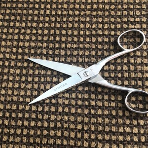 May include: Silver-toned metal sewing scissors featuring two symmetrical finger loops and sharp, pointed blades. The word SINGER is clearly engraved on one of the blades near the central pivot screw, which holds the two scissor halves together.