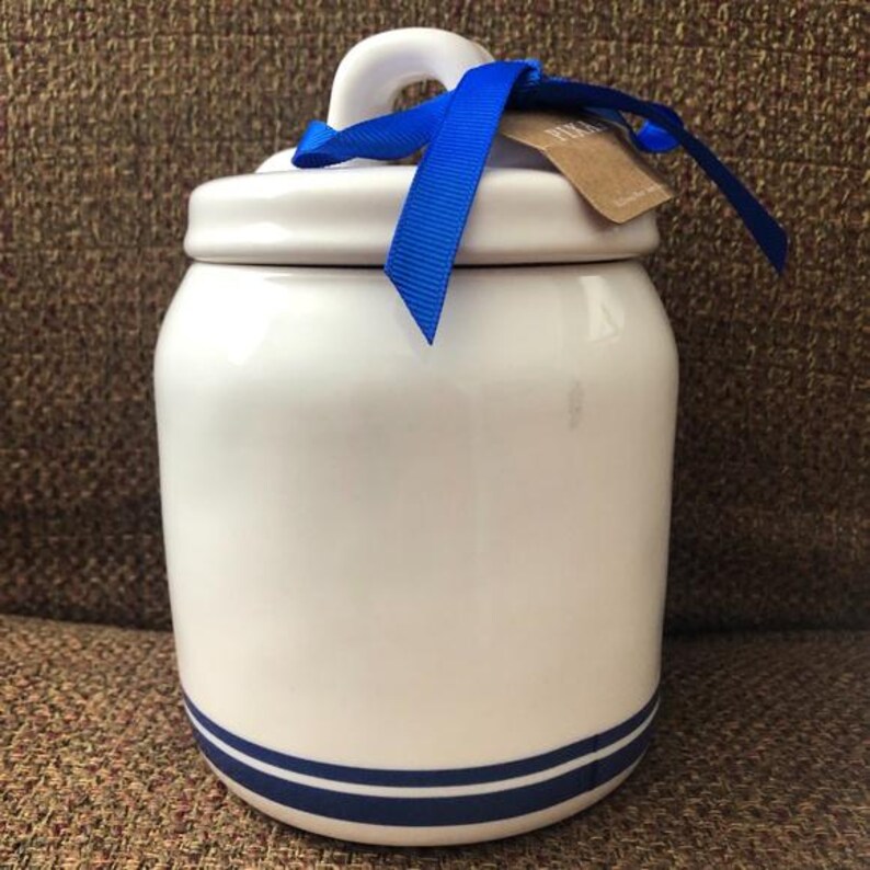 May include: White ceramic jar with a blue ribbon and a blue and white striped band around the bottom. The jar has a lid with a handle.