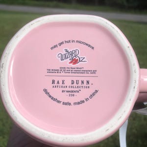 May include: The underside of a light pink ceramic mug featuring black printed text that reads The Wizard of Oz, Glinda the Good Witch, Rae Dunn Artisan Collection by Magenta, dishwasher safe, made in china, and may get hot in microwave.