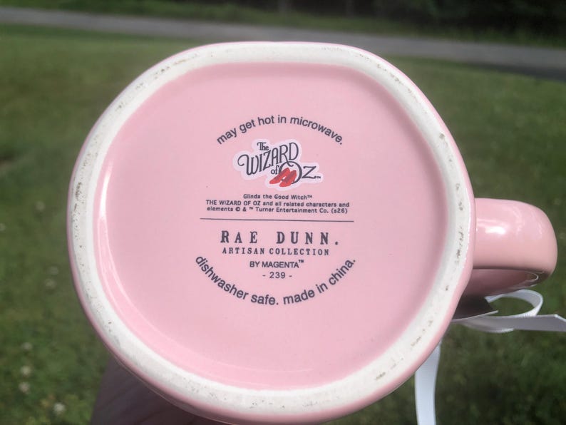 May include: The underside of a light pink ceramic mug featuring black printed text that reads The Wizard of Oz, Glinda the Good Witch, Rae Dunn Artisan Collection by Magenta, dishwasher safe, made in china, and may get hot in microwave.