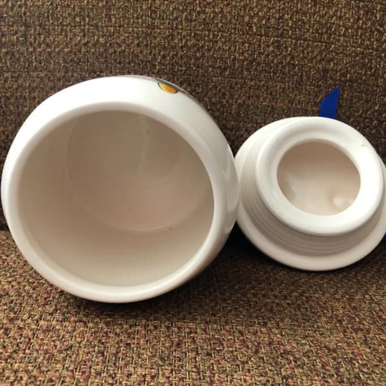 May include: White ceramic canister with a lid. The canister has a round shape and a smooth, white finish. The lid has a blue trim around the edge.