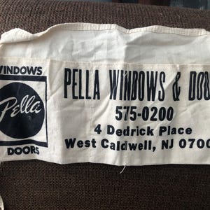 May include: Cream-colored fabric bag with black text. The bag reads "PELLA WINDOWS & DOORS" with a logo and contact information. The bag has a rectangular shape with a pocket and ties.