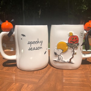 May include: Two white ceramic mugs with black handles. One mug has the words "spooky season" and bat graphics. The other mug features Snoopy with a pumpkin and a yellow moon. Both mugs have orange pumpkin decorations.