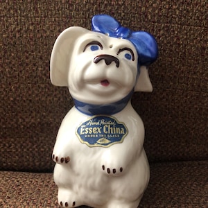 Puede incluir: Figurita de perro de cerámica blanca con un lazo y un collar azules. La figurita tiene ojos pintados de color marrón y una nariz negra. El perro está sentado con las patas metidas debajo del cuerpo. La figurita tiene una etiqueta azul y dorada que dice "Hand Painted Essex China Under The Glaze".