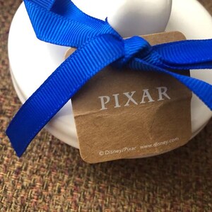 May include: A white ceramic figurine with a blue ribbon tied around it. The ribbon has a brown tag with the word "PIXAR" printed on it. The tag also includes the copyright information "&copy;Disney/Pixar www.disney.com".