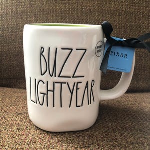 Puede incluir: Taza de cer&aacute;mica blanca con interior verde y texto negro que dice "Buzz Lightyear". Una etiqueta negra y azul con la palabra "PIXAR" est&aacute; unida al asa.