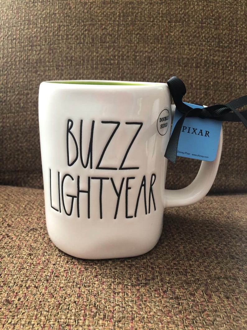 Puede incluir: Taza de cer&aacute;mica blanca con interior verde y texto negro que dice "Buzz Lightyear". Una etiqueta negra y azul con la palabra "PIXAR" est&aacute; unida al asa.