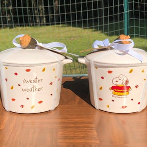 Puede incluir: Dos ollas de cerámica blanquecina con tapas, asas y decoraciones otoñales. Una olla tiene el texto "sweater weather" y la otra presenta un personaje de dibujos animados. Ambas están atadas con cintas blancas.