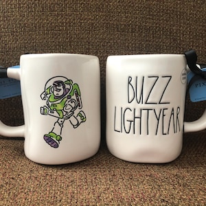 Puede incluir: Dos tazas de cer&aacute;mica blanca con asas negras. Una taza presenta una imagen de dibujos animados de Buzz Lightyear de la franquicia Toy Story. La otra taza tiene el texto "Buzz Lightyear" en letras negras. Ambas tazas tienen una etiqueta azul y blanca adjunta.