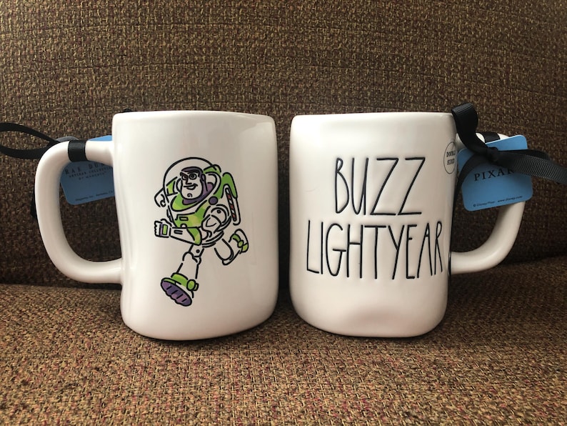 Puede incluir: Dos tazas de cer&aacute;mica blanca con asas negras. Una taza presenta una imagen de dibujos animados de Buzz Lightyear de la franquicia Toy Story. La otra taza tiene el texto "Buzz Lightyear" en letras negras. Ambas tazas tienen una etiqueta azul y blanca adjunta.